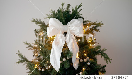 large white bow on the top of an evergreen tree, decorated with lights and garland, tied in the front with thick fabric large white bow on the top of an evergreen tree, decorated with lights and garland, tied in the front with thick fabric 137104875