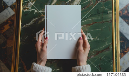 person holding up an empty white book mockup, sitting at a table with their hands on it. the background is green and gold, creating a luxurious atmosphere person holding up an empty white book mockup, sitting at a table with their hands on it. the background is green and gold, creating a luxurious atmosphere 137104883