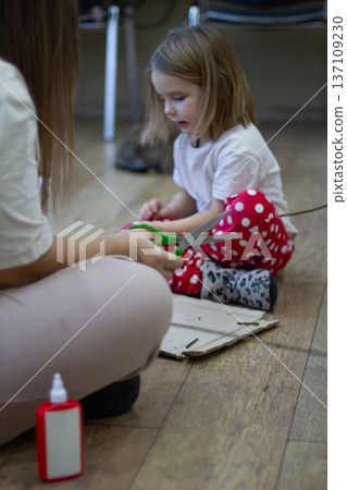 Vertical. A mother and daughter are making a homemade toy out of cardboard, branches, and natural materials, cutting and gluing the pieces together. The family is enjoying spending time together. 137109230
