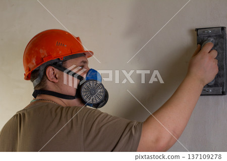 A construction worker wearing personal protective equipment, including a hard hat and a respirator, sands the plaster of an interior wall. Leveling the wall with a sanding machine. A construction worker wearing personal protective equipment, including a hard hat and a respirator, sands the plaster of an interior wall. Leveling the wall with a sanding machine. 137109278