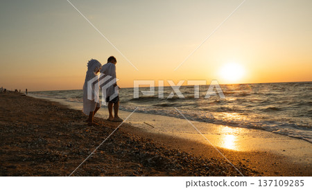 Two children, one wearing a hooded towel, standing by the water's edge on a pebble beach, watching the setting sun over the ocean waves during a summer vacation. Two children, one wearing a hooded towel, standing by the water's edge on a pebble beach, watching the setting sun over the ocean waves during a summer vacation. 137109285