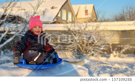 Cheerful young child wearing a pink hat and winter clothing, sitting on a blue sled, laughing and playing on a snowy outdoor day with houses and bare trees in background. 137109288