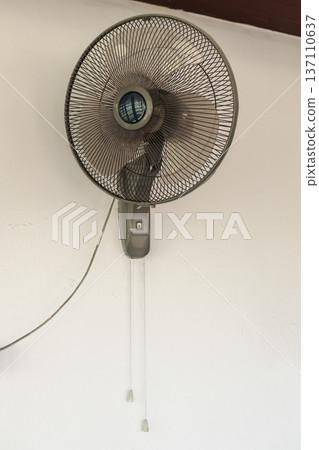 Wall mounted electric fan with pull cords on white wall providing ventilation and cooling in tropical house interior 137110637