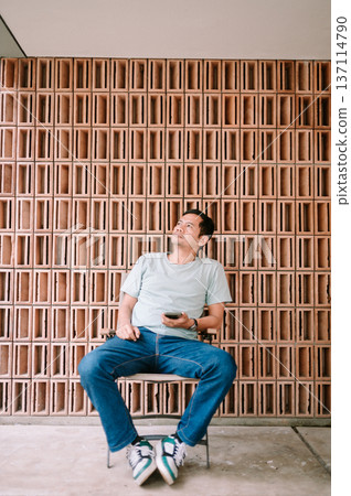 Thoughtful Man Holding Phone Sitting by Architectural Terracotta Wall 137114790
