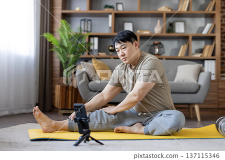 Asian man on a yellow yoga mat following an online fitness routine on his smartphone at home, stretching and exercising in the living room for wellness and self-care Asian man on a yellow yoga mat following an online fitness routine on his smartphone at home, stretching and exercising in the living room for wellness and self-care 137115346