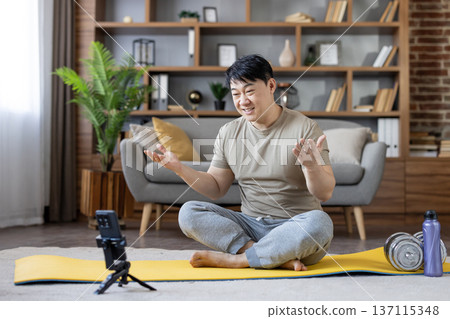 Asian man sitting on a yellow yoga mat, streaming a fitness tutorial or live workout session from his living room, actively gesturing while filming on a smartphone Asian man sitting on a yellow yoga mat, streaming a fitness tutorial or live workout session from his living room, actively gesturing while filming on a smartphone 137115348
