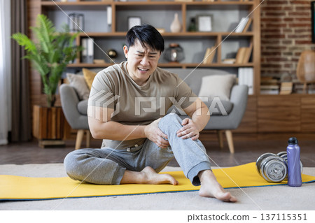 Asian man sitting on yoga mat at home clutching his knee with a pained expression after exercise, depicting knee pain, strain or injury during a workout or stretching session Asian man sitting on yoga mat at home clutching his knee with a pained expression after exercise, depicting knee pain, strain or injury during a workout or stretching session 137115351