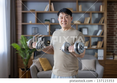 Asian man smiling while doing bicep curls with dumbbells in his living room, focusing on strength training and maintaining a healthy lifestyle through home workouts Asian man smiling while doing bicep curls with dumbbells in his living room, focusing on strength training and maintaining a healthy lifestyle through home workouts 137115353