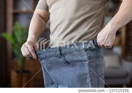 Man demonstrating successful weight loss and health transformation, holding out the waist of his loose, oversized jeans, symbolizing achievement in diet and fitness 137115356