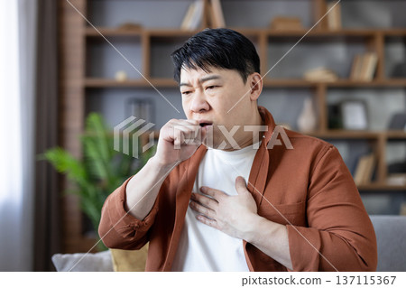Asian man at home clutching chest and covering his mouth while coughing, showing flu-like symptoms and discomfort as he rests in a living room during illness and recovery Asian man at home clutching chest and covering his mouth while coughing, showing flu-like symptoms and discomfort as he rests in a living room during illness and recovery 137115367