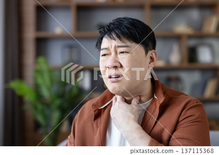Asian man at home with painful sore throat, touching his neck and grimacing from illness and discomfort, appearing unwell and fatigued while suffering respiratory symptoms Asian man at home with painful sore throat, touching his neck and grimacing from illness and discomfort, appearing unwell and fatigued while suffering respiratory symptoms 137115368