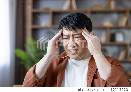 Middle-aged asian man experiencing intense headache or migraine, grimacing and touching his temples in an attempt to alleviate the discomfort while feeling stressed at home Middle-aged asian man experiencing intense headache or migraine, grimacing and touching his temples in an attempt to alleviate the discomfort while feeling stressed at home 137115370