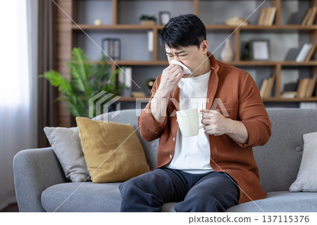 Young asian man sitting on a sofa in his living room, suffering from a cold, flu, or allergy, blowing his nose with a tissue while holding a warm mug for comfort Young asian man sitting on a sofa in his living room, suffering from a cold, flu, or allergy, blowing his nose with a tissue while holding a warm mug for comfort 137115376