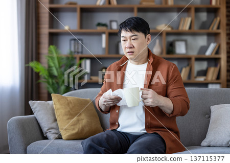 Asian man holding a tissue and a warm drink while sitting on a couch at home, experiencing cold or flu symptoms and feeling sick, needing to rest and recover 137115377