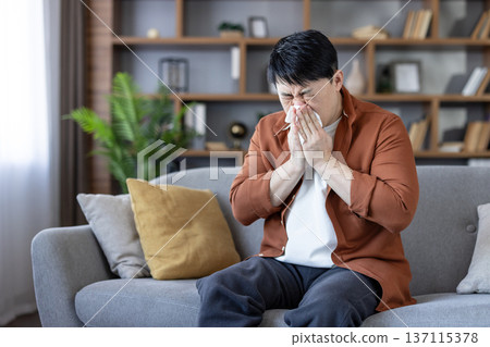 Asian man sitting on a couch at home, suffering from an illness, allergy, or cold, covering his nose with a tissue while sneezing and showing discomfort Asian man sitting on a couch at home, suffering from an illness, allergy, or cold, covering his nose with a tissue while sneezing and showing discomfort 137115378