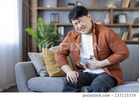 Asian man sitting on sofa at home clutching his abdomen in pain, showing discomfort from stomach ache, indigestion or cramp, unwell and frustrated in living room Asian man sitting on sofa at home clutching his abdomen in pain, showing discomfort from stomach ache, indigestion or cramp, unwell and frustrated in living room 137115379