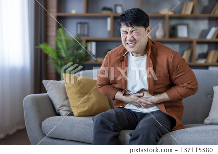Man clutching his abdomen and grimacing on a living room couch, suffering from severe stomach pain and discomfort from indigestion or other gastrointestinal illness at home Man clutching his abdomen and grimacing on a living room couch, suffering from severe stomach pain and discomfort from indigestion or other gastrointestinal illness at home 137115380