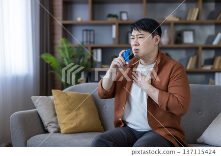 Asian man sitting on a sofa at home using a blue inhaler, experiencing shortness of breath and difficulty breathing during an asthma attack, holding a hand to his chest Asian man sitting on a sofa at home using a blue inhaler, experiencing shortness of breath and difficulty breathing during an asthma attack, holding a hand to his chest 137115424