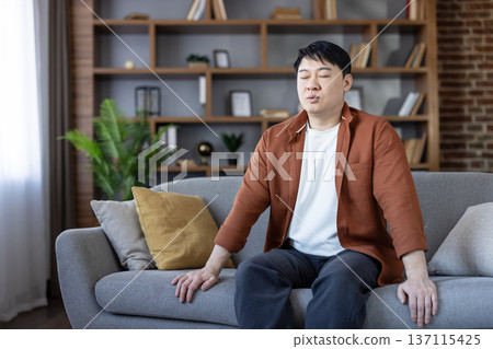 Asian man sitting relaxed on a comfortable sofa with closed eyes, engaging in a mindful breathing exercise or meditation for stress relief and mental well-being in his living room Asian man sitting relaxed on a comfortable sofa with closed eyes, engaging in a mindful breathing exercise or meditation for stress relief and mental well-being in his living room 137115425