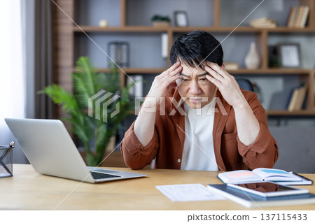 Asian man at home office clutching his head in frustration, exhausted and overwhelmed while working on a laptop, showing stress, headache and burnout during remote work 137115433