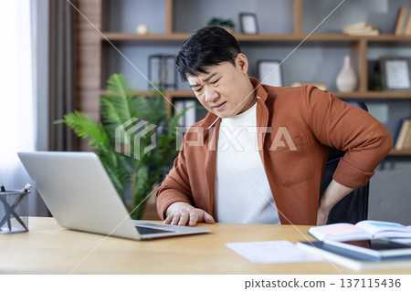 Asian man experiencing severe lower back pain while sitting at a desk and working on his laptop in a home office setting, indicating health issues from prolonged sitting Asian man experiencing severe lower back pain while sitting at a desk and working on his laptop in a home office setting, indicating health issues from prolonged sitting 137115436