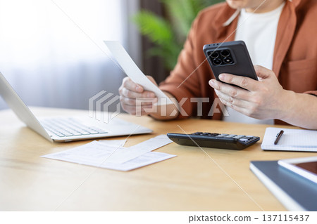 Person calculating financial budget, analyzing invoices with a mobile application, managing personal finance, and planning household expenses on a wooden desk Person calculating financial budget, analyzing invoices with a mobile application, managing personal finance, and planning household expenses on a wooden desk 137115437