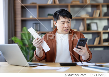 Asian man sitting at a desk and reviewing unexpected high bills, holding a phone and a paper document, reacting with a shocked and stressed expression about rising prices and financial challenges 137115439