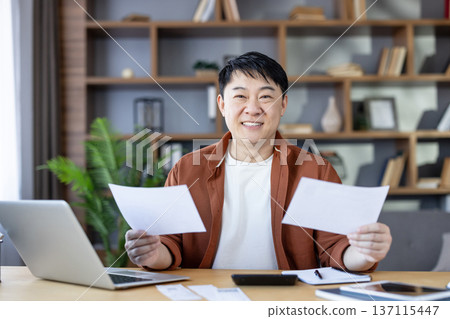 Asian man sitting at desk in home office, holding papers and looking at camera, actively managing personal finances and household budget with a laptop, feeling successful 137115447