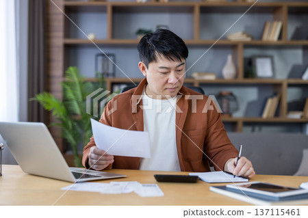 Asian man sitting at a desk in his home office, reviewing financial documents and bills, while writing notes in a notebook next to a laptop and calculator, focused on managing his personal budget 137115461