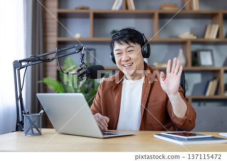Smiling asian man wearing headphones and waving while recording a podcast or live streaming content using a microphone and laptop from his home office Smiling asian man wearing headphones and waving while recording a podcast or live streaming content using a microphone and laptop from his home office 137115472