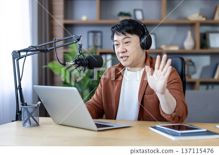 Asian man with headphones speaking into professional microphone, waving hand while recording online podcast or broadcasting live from home studio using a laptop 137115476