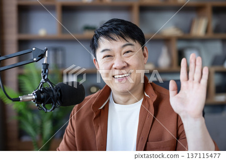 Asian man creating content, smiling and waving, broadcasting a live stream or recording a podcast in a home studio, connecting with his online audience 137115477