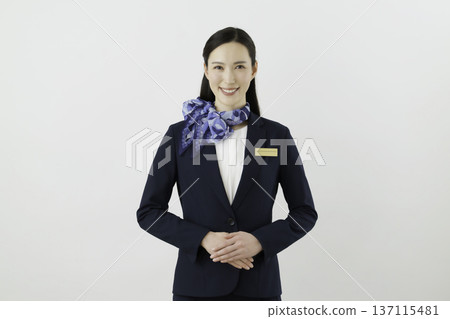 一位面帶微笑的日本女門房，身穿藏藍色套裝，佩戴絲帶和名牌，背景為白色。 137115481