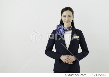 一位面帶微笑的日本女門房，身穿藏藍色套裝，佩戴絲帶和名牌，背景為白色。 137115482
