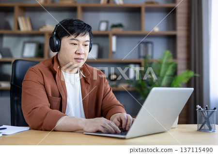 Asian man with headphones concentrating on his laptop, typing and working remotely from a modern home office setting during the day, showing dedication to his online tasks 137115490
