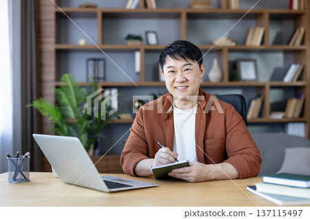 Smiling asian man sitting at a desk with a laptop, holding a small notebook and pen, working from his home office, demonstrating remote work and productivity Smiling asian man sitting at a desk with a laptop, holding a small notebook and pen, working from his home office, demonstrating remote work and productivity 137115497