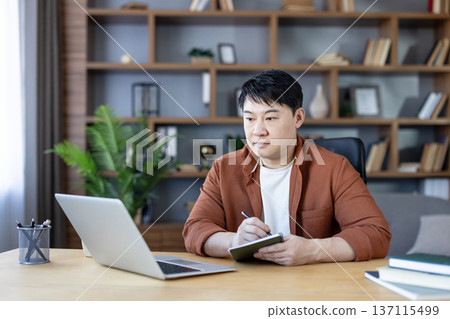 Attentive asian man focusing on a laptop screen while writing in a notebook, working or studying at a wooden desk in a modern home office with bookshelves Attentive asian man focusing on a laptop screen while writing in a notebook, working or studying at a wooden desk in a modern home office with bookshelves 137115499