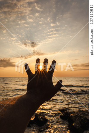 Silhouette hand holding sun over sea horizon at sunset with dramatic sky background Silhouette hand holding sun over sea horizon at sunset with dramatic sky background 137115912
