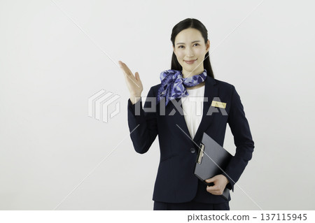 一位面帶微笑的日本女門房,身穿藏藍色套裝,佩戴絲帶和名牌,在白色背景前舉起手。 一位面帶微笑的日本女門房,身穿藏藍色套裝,佩戴絲帶和名牌,在白色背景前舉起手。 137115945