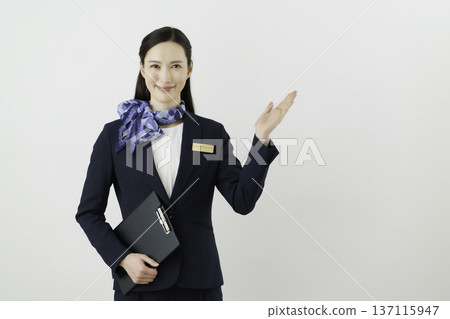 一位面帶微笑的日本女門房，身穿藏藍色套裝，佩戴絲帶和名牌，在白色背景前舉起手。 137115947