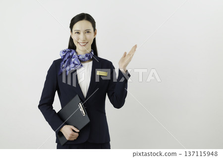 一位面帶微笑的日本女門房，身穿藏藍色套裝，佩戴絲帶和名牌，在白色背景前舉起手。 137115948