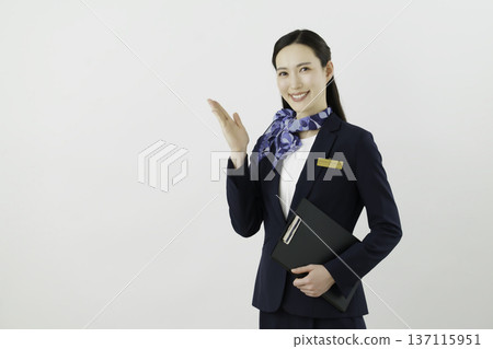一位面帶微笑的日本女門房，身穿藏藍色套裝，佩戴絲帶和名牌，在白色背景前舉起手。 137115951