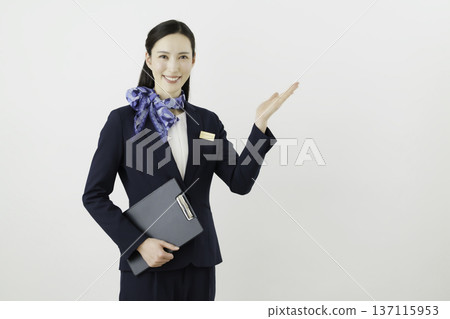 一位面帶微笑的日本女門房，身穿藏藍色套裝，佩戴絲帶和名牌，在白色背景前舉起手。 137115953