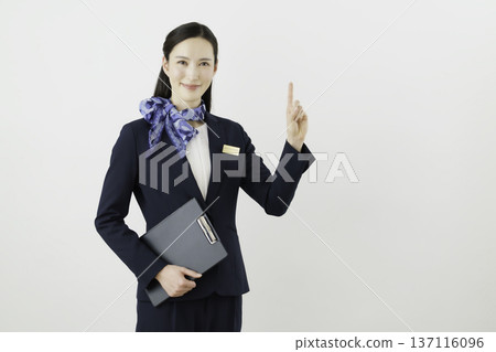 一位面帶微笑的日本女門房，身穿藏藍色套裝，佩戴絲帶和名牌，在白色背景下擺出“1號”的姿勢。 137116096