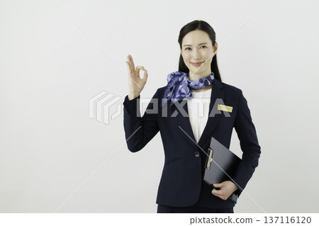 一位面帶微笑的日本女性，身穿藏藍色套裝，佩戴絲帶和名牌，站在白色背景前，手指比出一個“OK”的手勢。禮賓部 137116120