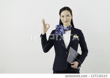 一位面帶微笑的日本女性，身穿藏藍色套裝，佩戴絲帶和名牌，站在白色背景前，手指比出一個“OK”的手勢。禮賓部 137116123