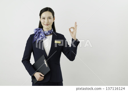 一位面帶微笑的日本女性，身穿藏藍色套裝，佩戴絲帶和名牌，站在白色背景前，手指比出一個“OK”的手勢。禮賓部 137116124