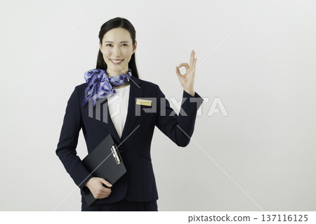 A smiling Japanese woman wearing a navy suit, ribbon, and name tag on a white background, making a circle OK pose with her fingers. Concierge 137116125