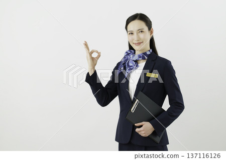 一位面帶微笑的日本女性,身穿藏藍色套裝,佩戴絲帶和名牌,站在白色背景前,手指比出一個“OK”的手勢。禮賓部 一位面帶微笑的日本女性,身穿藏藍色套裝,佩戴絲帶和名牌,站在白色背景前,手指比出一個“OK”的手勢。禮賓部 137116126