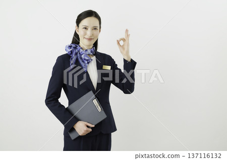 一位面帶微笑的日本女性，身穿藏藍色套裝，佩戴絲帶和名牌，站在白色背景前，手指比出一個“OK”的手勢。禮賓部 137116132